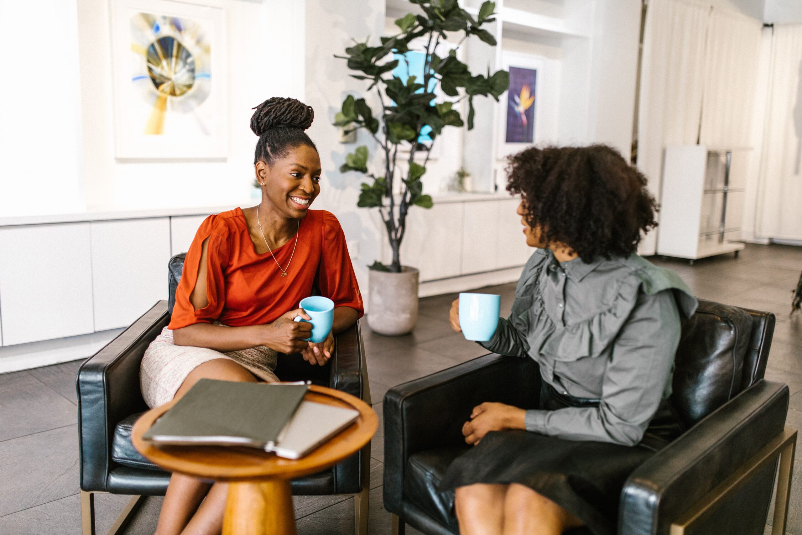Two women sitting in mid-conversation with smiles, holding coffee cups in an office setting.